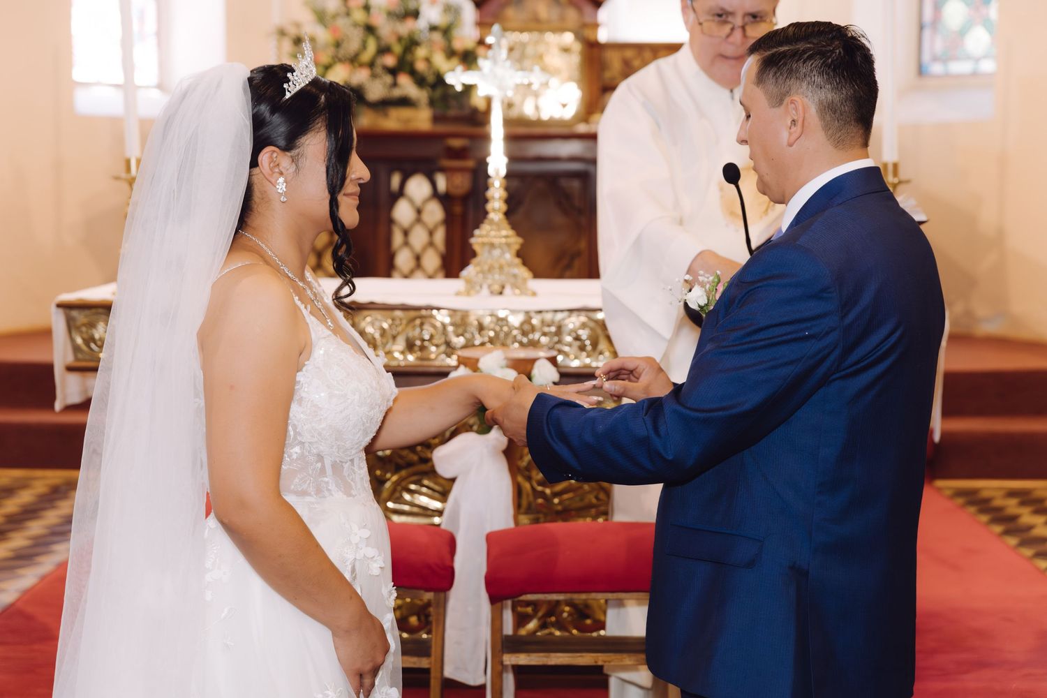 Groom in blue suit exchanges rings with bride in white gown during church wedding ceremony.
