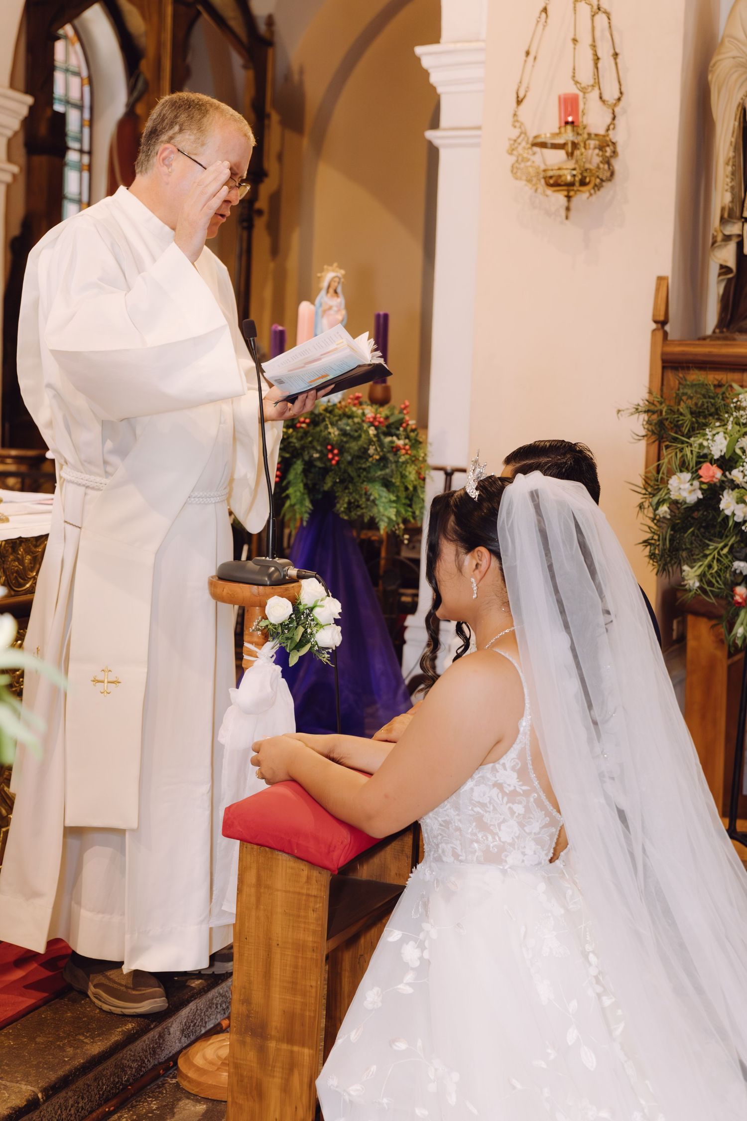 Priest reads scripture as bride in white lace gown kneels during church wedding ceremony.