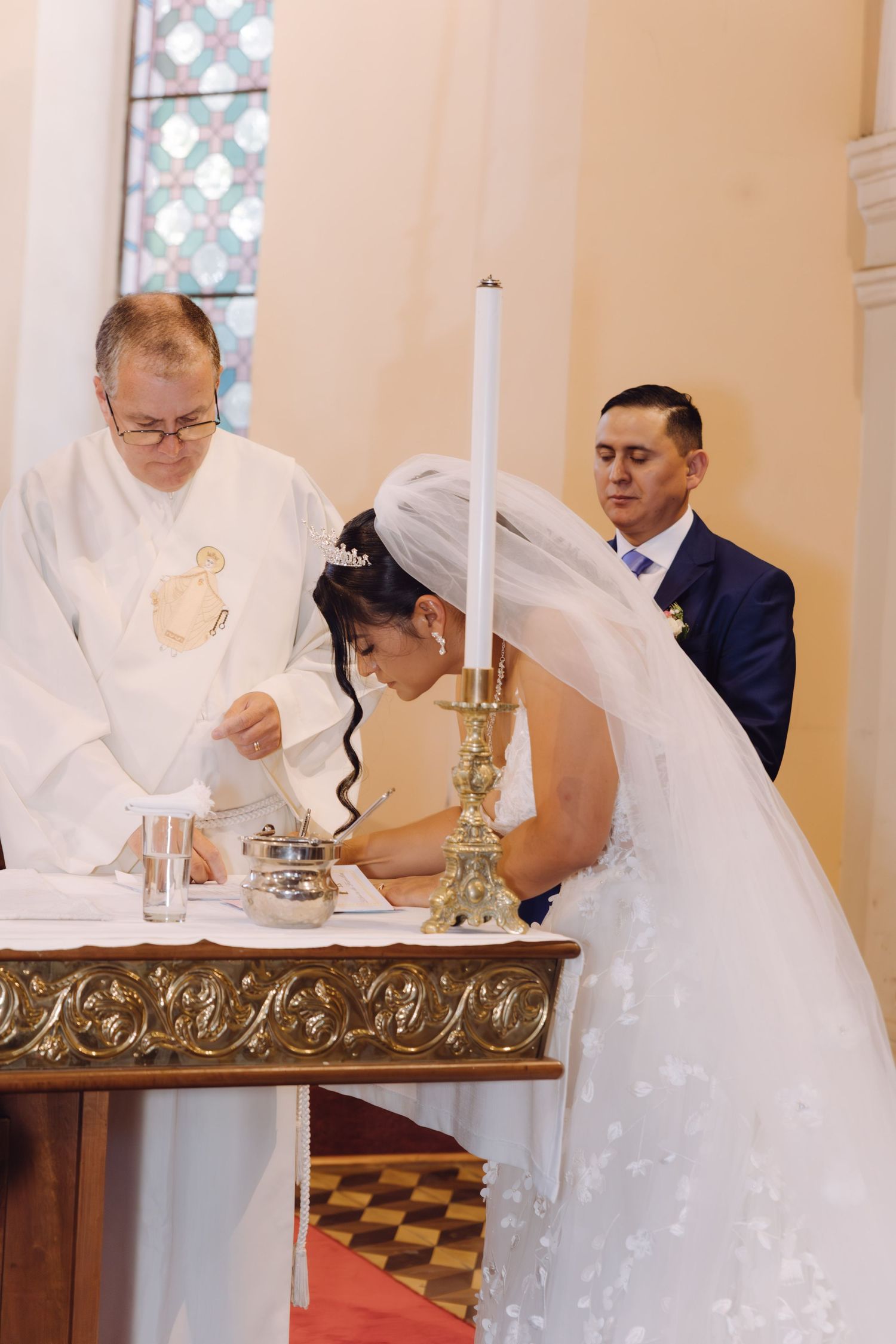 Bride in white veil signs marriage register at ornate altar with priest and groom nearby.
