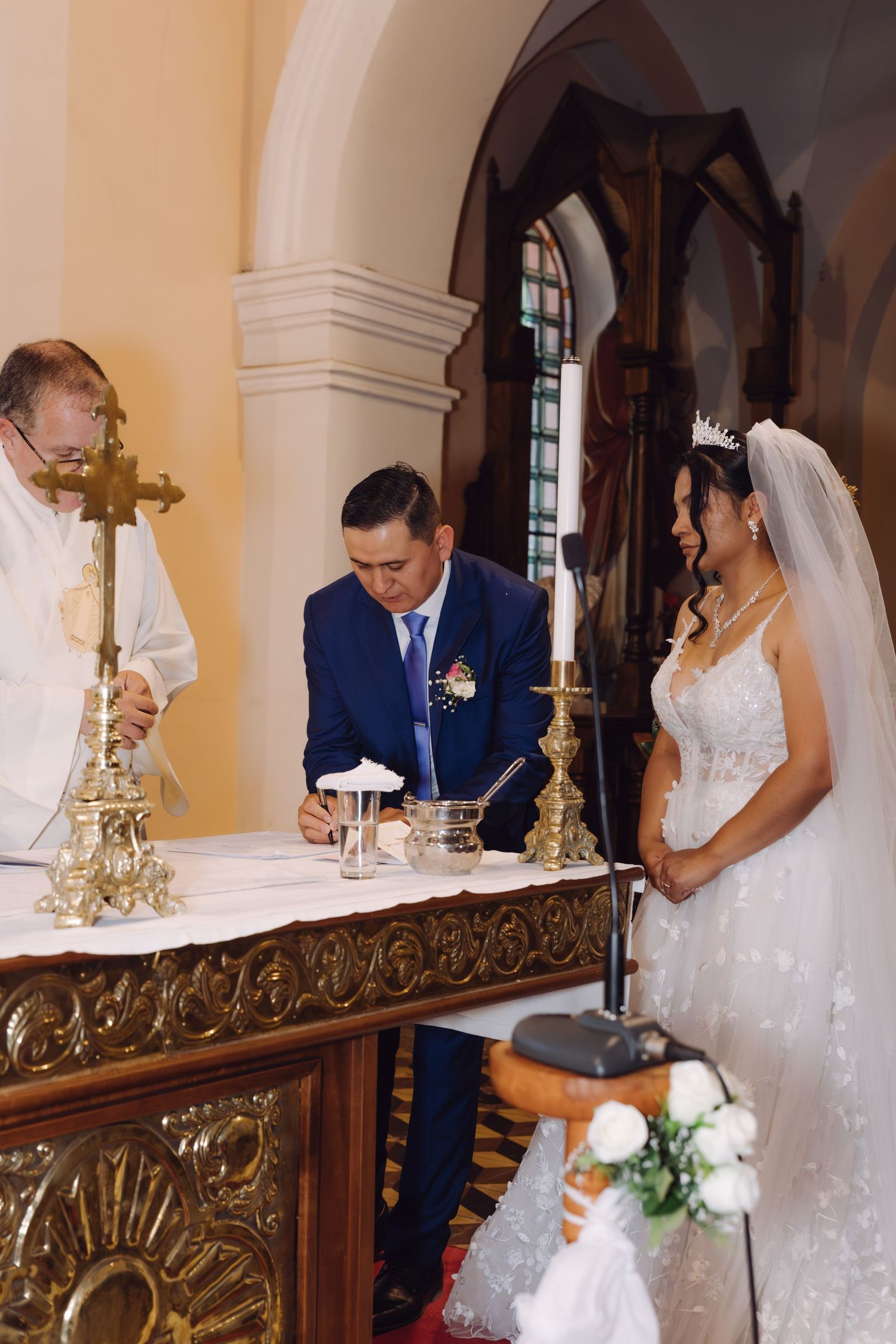 Groom in blue suit signs wedding register at church altar while bride in tiara looks on.