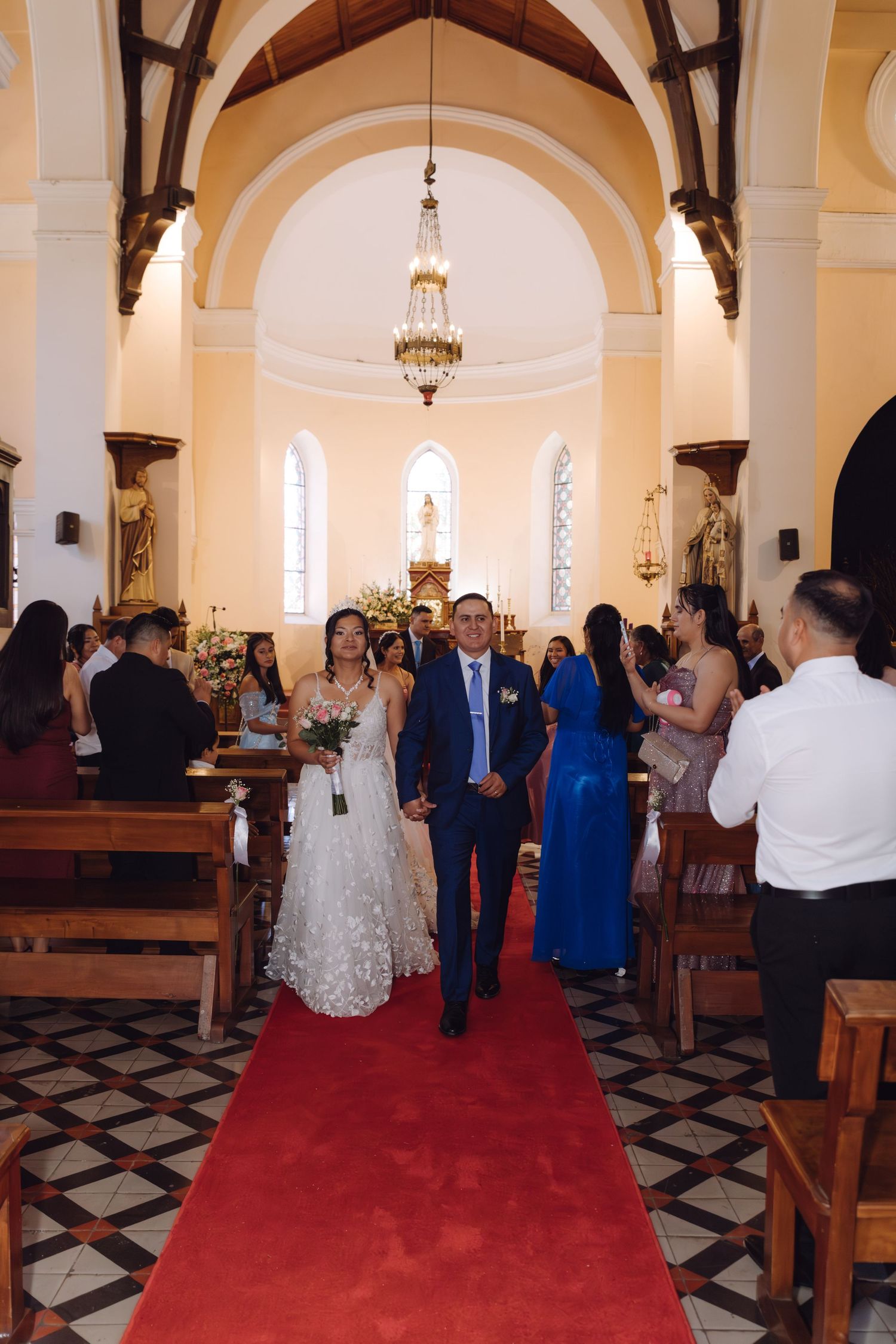 Newlyweds walk down red carpet aisle as guests applaud in elegant Catholic church.