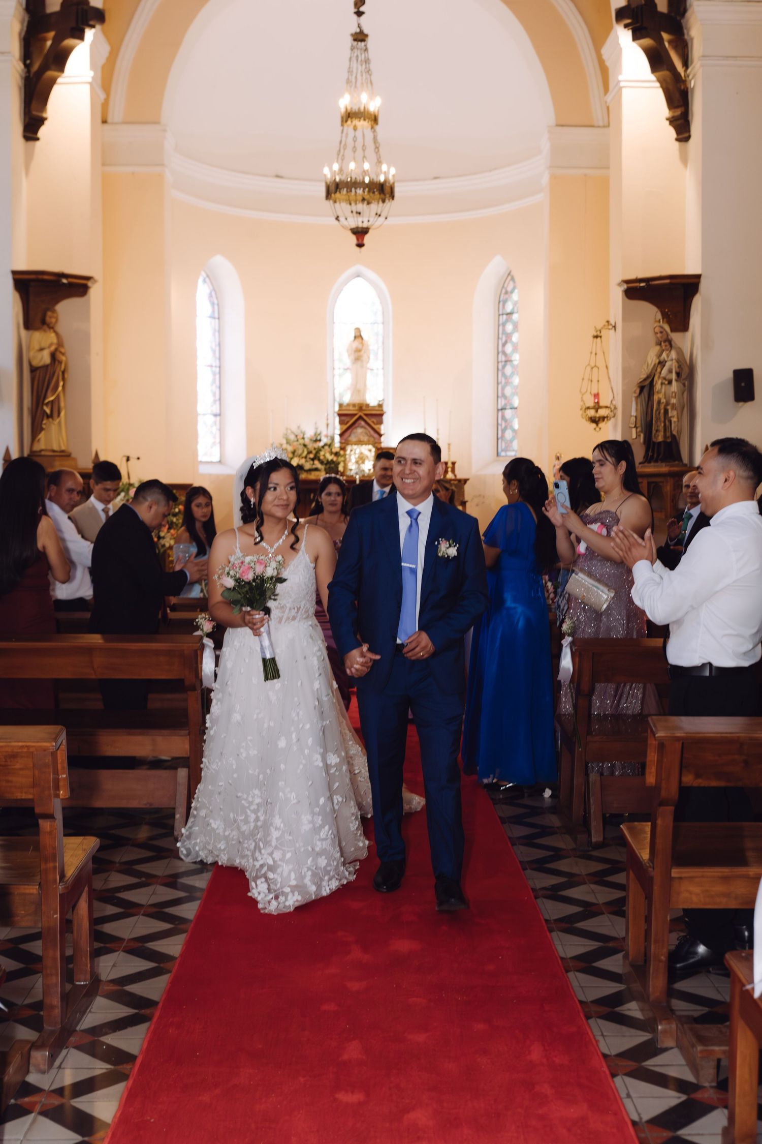 Smiling newlyweds walk hand in hand down red aisle as guests celebrate in church.