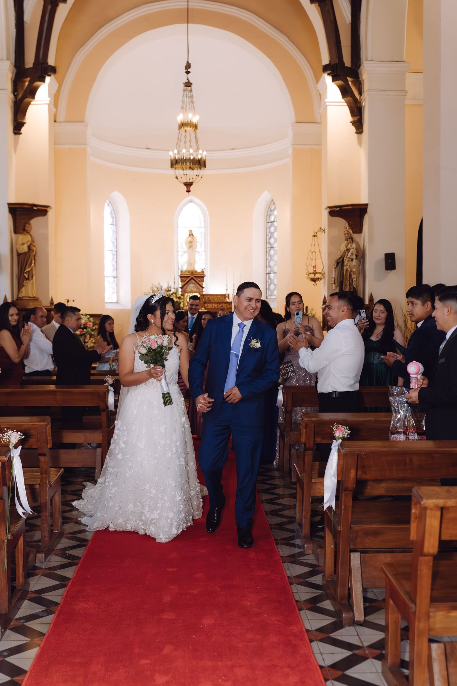 Bride and groom walk joyfully down red carpet aisle surrounded by cheering wedding guests.