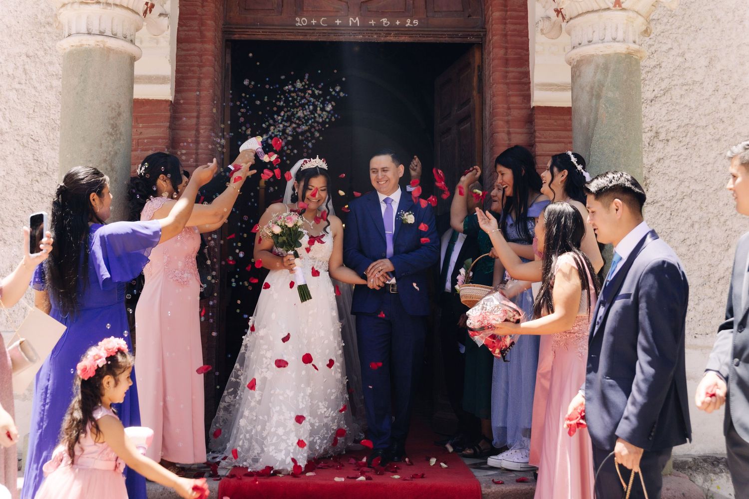 Wedding couple walks through shower of rose petals thrown by smiling guests outside church.