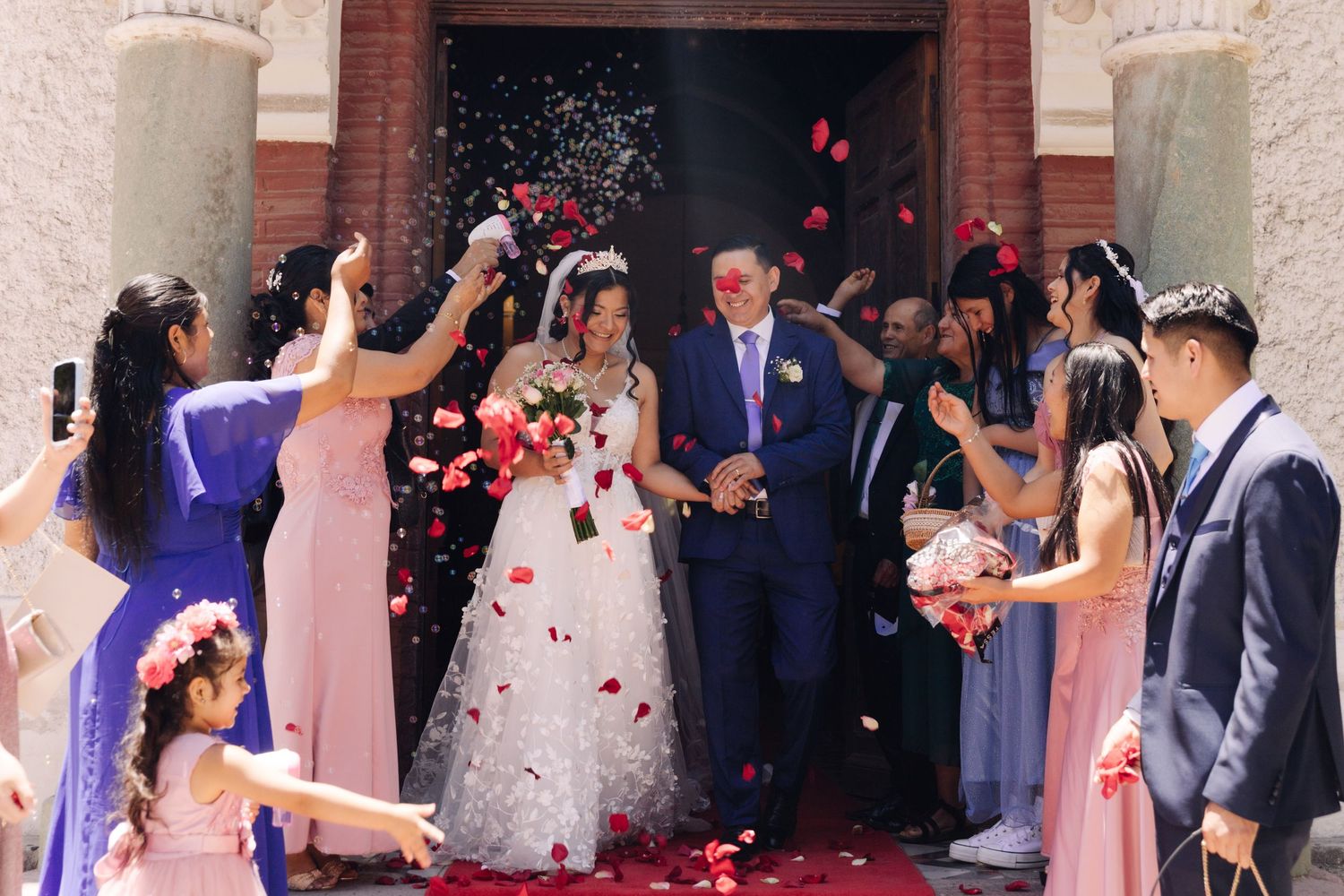 Newlyweds leave church entrance amid colorful confetti and rose petals from celebrating guests.