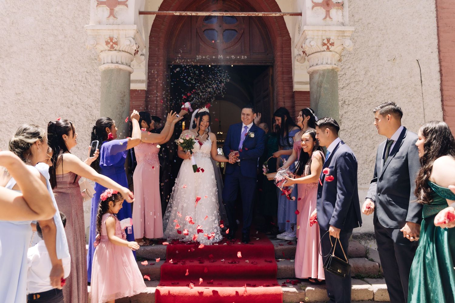 Bride and groom descend red carpet steps as guests shower them with rose petals.