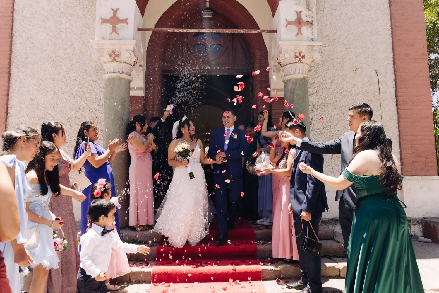 Joyful wedding party tosses confetti and petals as newlyweds exit decorated church doors.