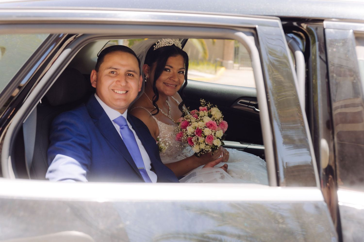 Smiling newlyweds sit together in wedding car, bride holding pink and white bouquet.