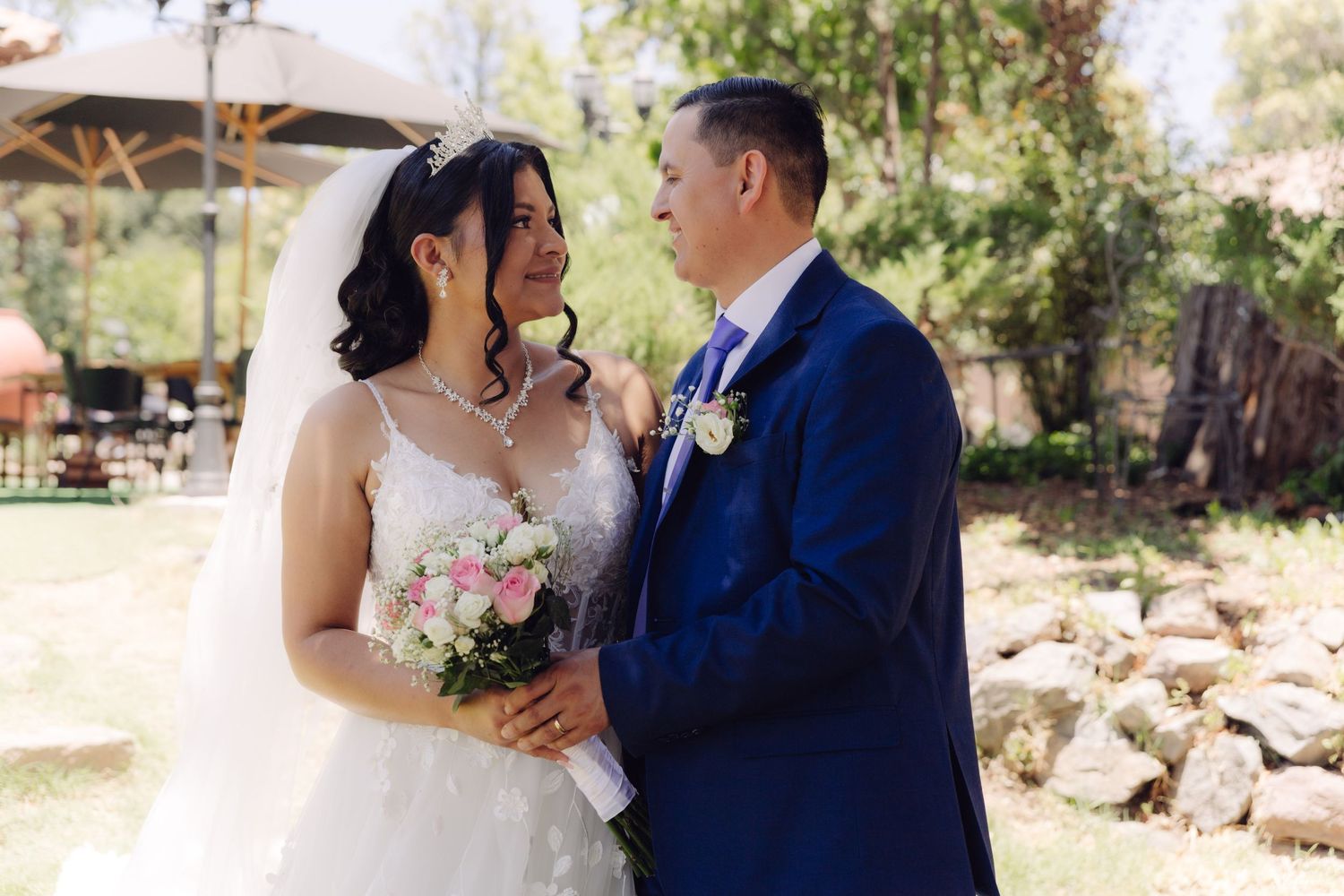 Bride and groom gaze at each other lovingly outdoors, bride holding elegant floral bouquet.