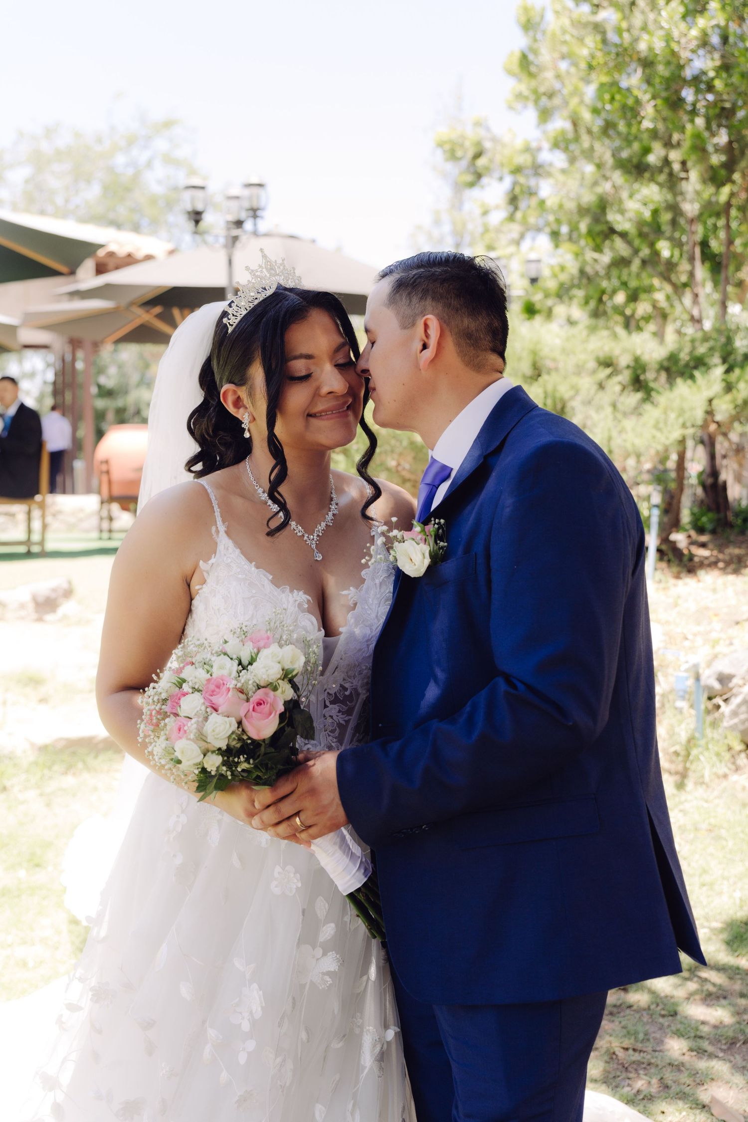 Groom in blue suit kisses bride wearing white lace gown and tiara while she holds pink bouquet outdoors.