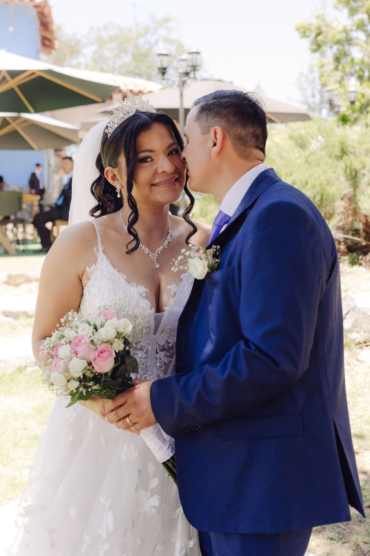 Smiling bride in white lace dress and tiara holds pink bouquet as groom in blue suit whispers to her.