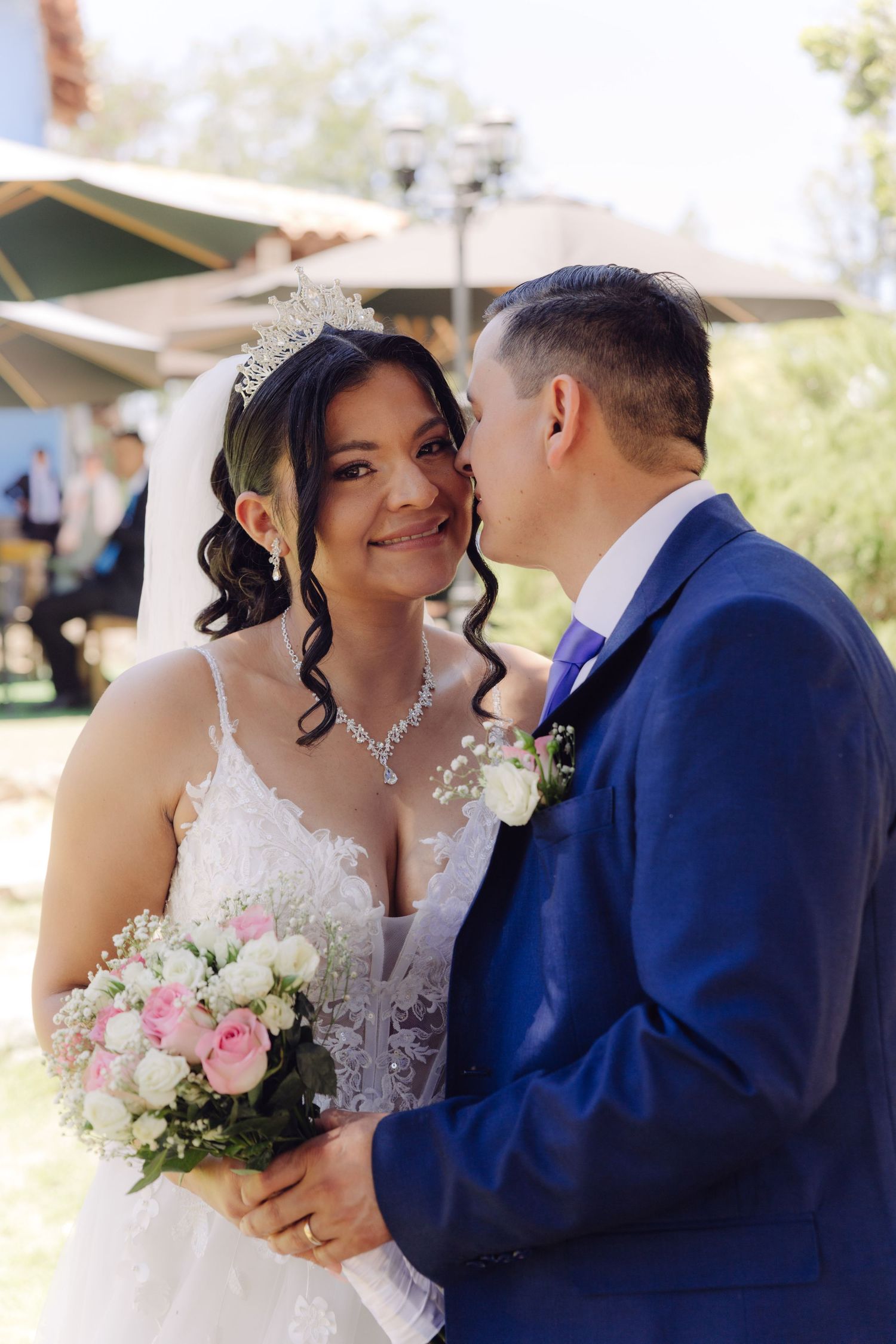 Bride in white lace gown and crystal tiara smiles as groom in navy suit kisses her cheek outdoors.