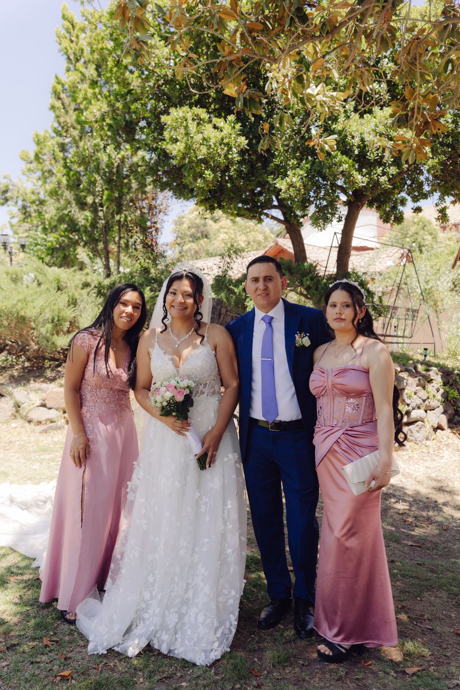 Wedding couple in white gown and blue suit pose with two bridesmaids wearing pink dresses outdoors.