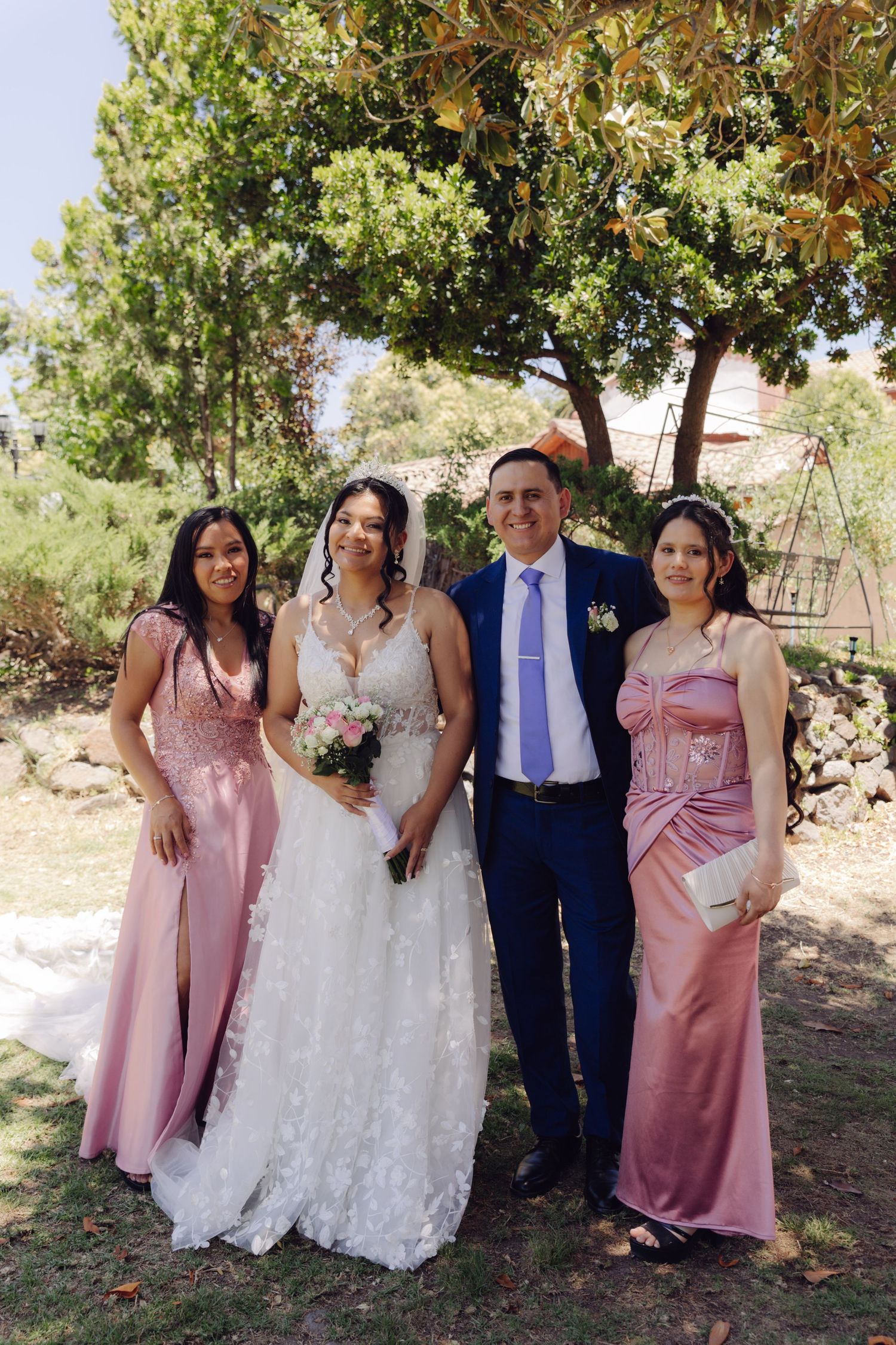 Bride in white gown and groom in blue suit pose with two bridesmaids in pink dresses under trees.