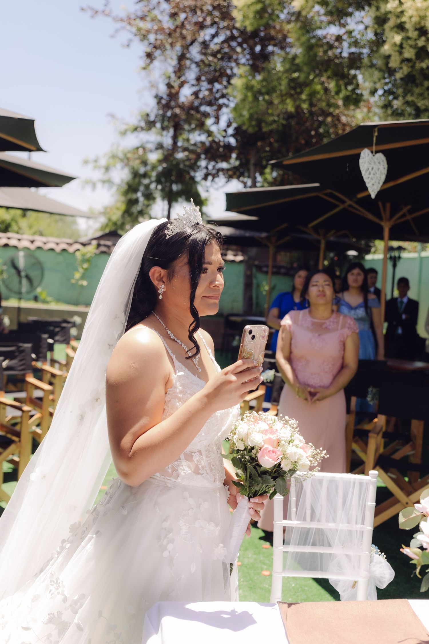 Bride in white lace wedding dress holds bouquet and phone during outdoor wedding ceremony with guests.