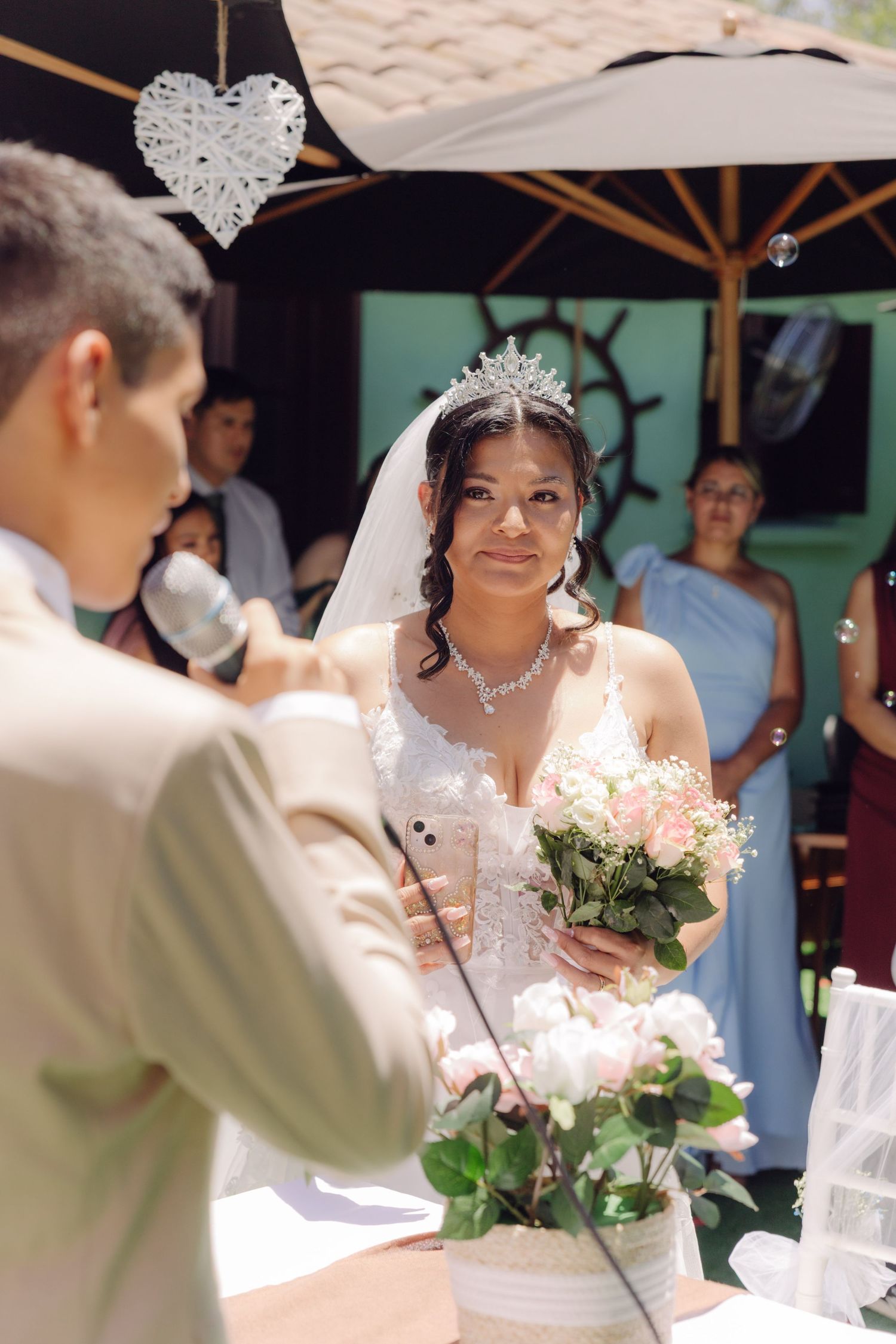 Bride in white lace gown and crystal tiara holds pink bouquet listening to speaker at outdoor ceremony.