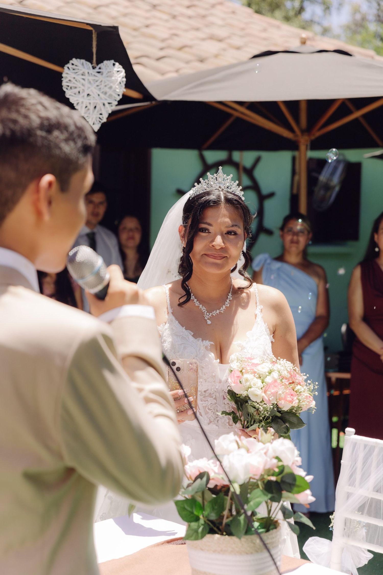 Bride wearing white lace dress and tiara holds pink bouquet while listening at outdoor wedding ceremony.