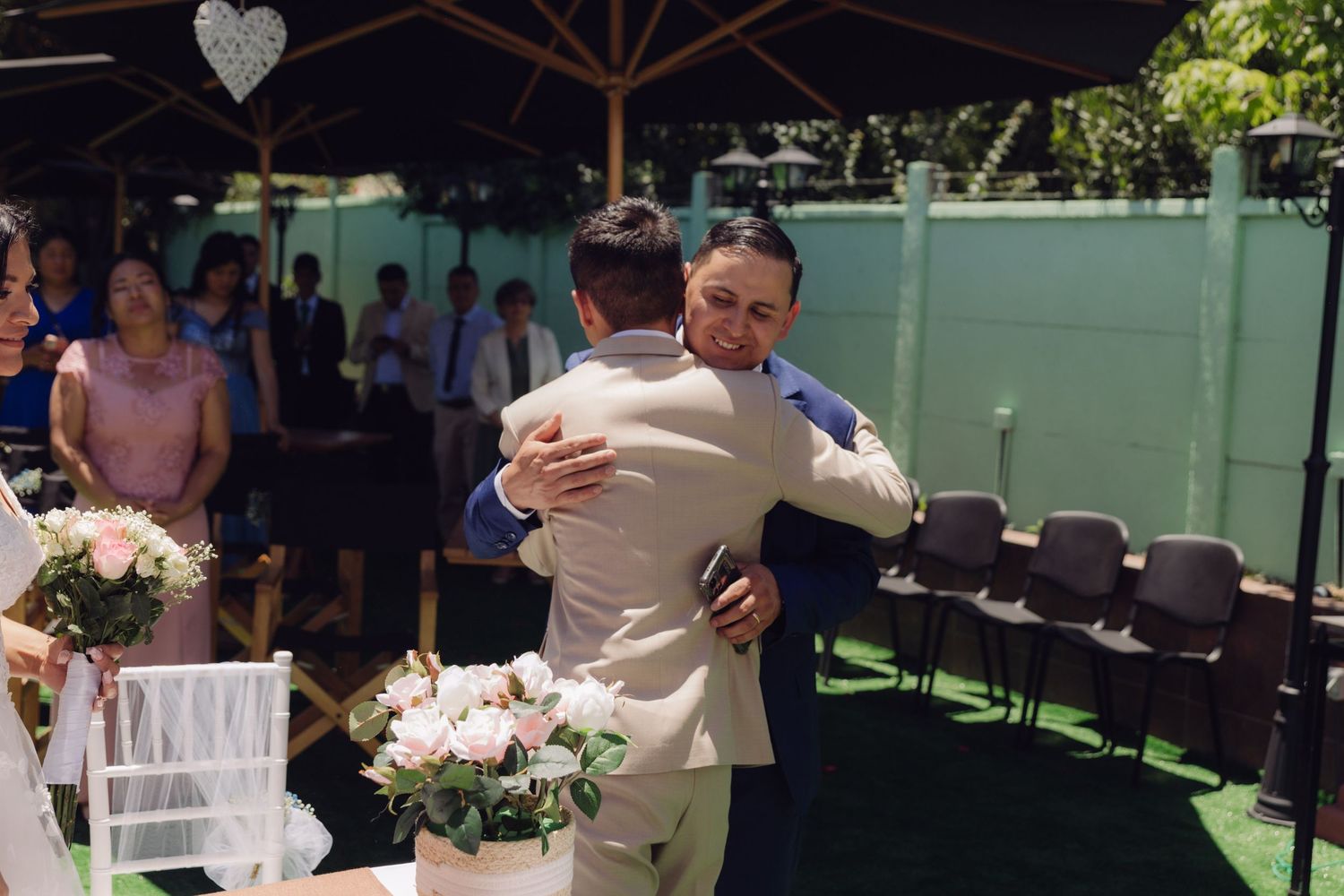 Bride in white wedding dress hugs a guest in beige suit at outdoor wedding reception venue.