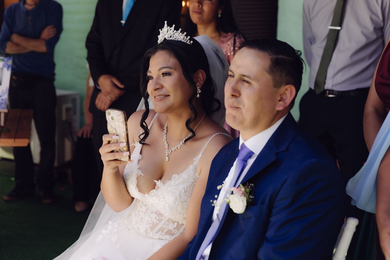 Bride wearing tiara and white gown looks at phone while groom in blue suit sits beside her at outdoor wedding.