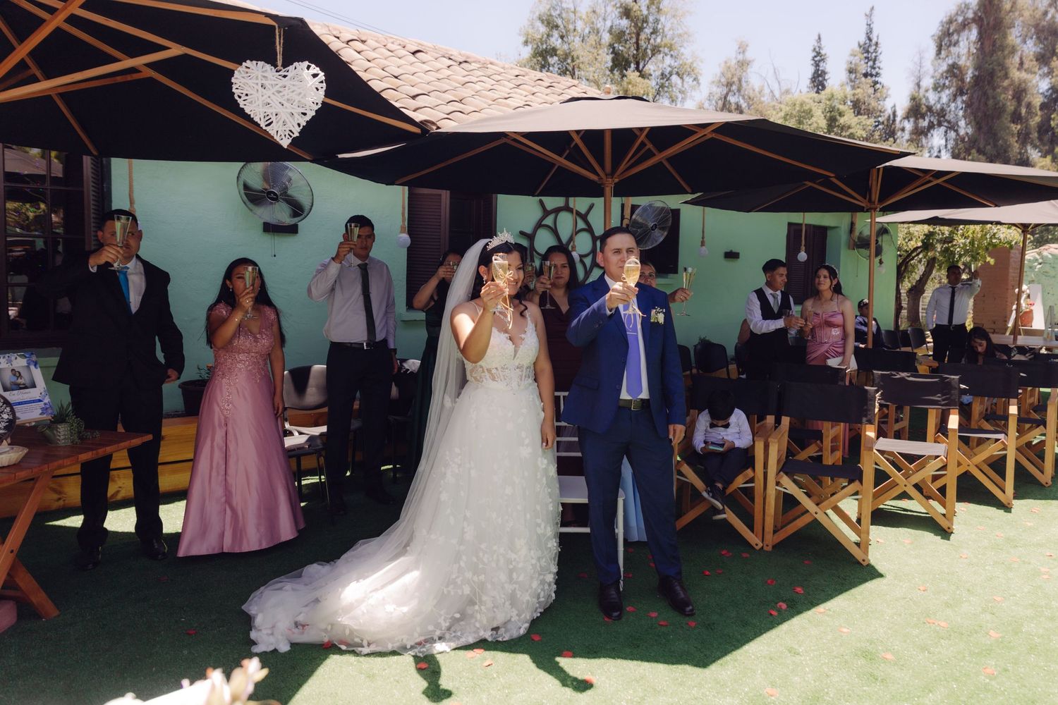 Bride in lace gown and groom in blue suit stand together at outdoor wedding venue with guests seated.