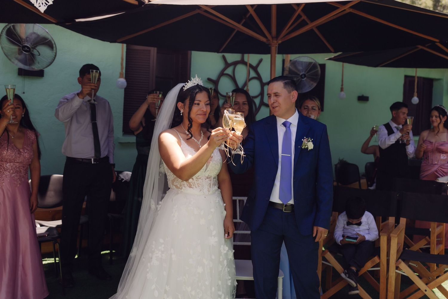 Bride and groom raise champagne glasses in toast at outdoor wedding reception under umbrella.