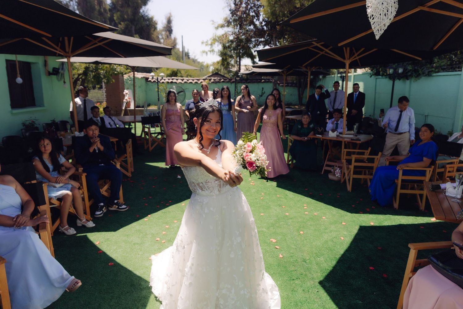 Bride in white strapless gown tosses bouquet to guests at outdoor wedding reception on green lawn.