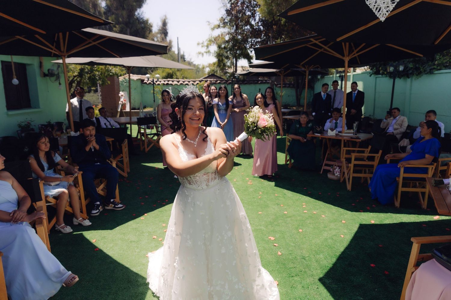 Bride in white gown prepares to toss bouquet to guests seated outdoors at wedding reception.