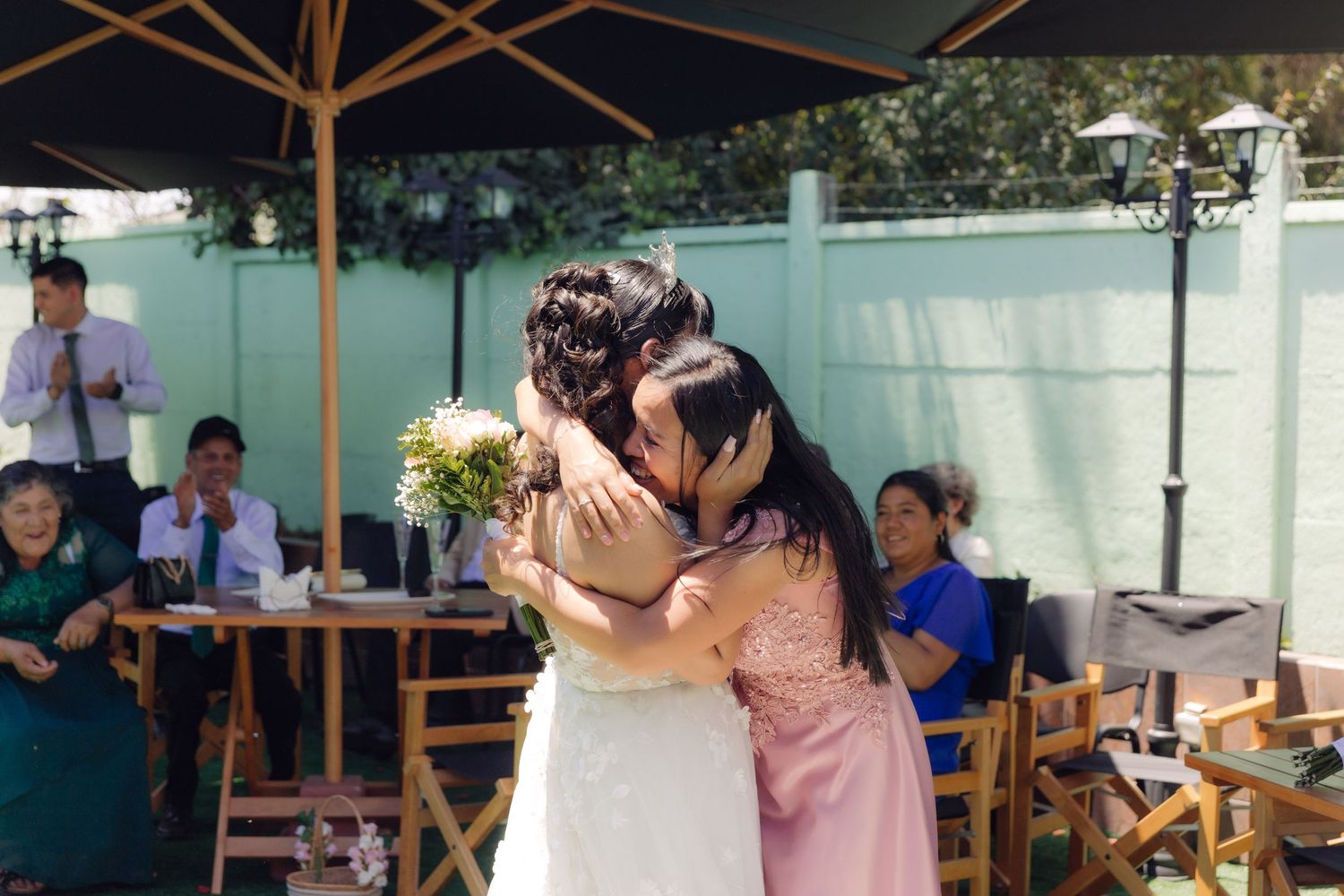 Bride and guest share emotional hug at outdoor wedding reception under patio umbrella.