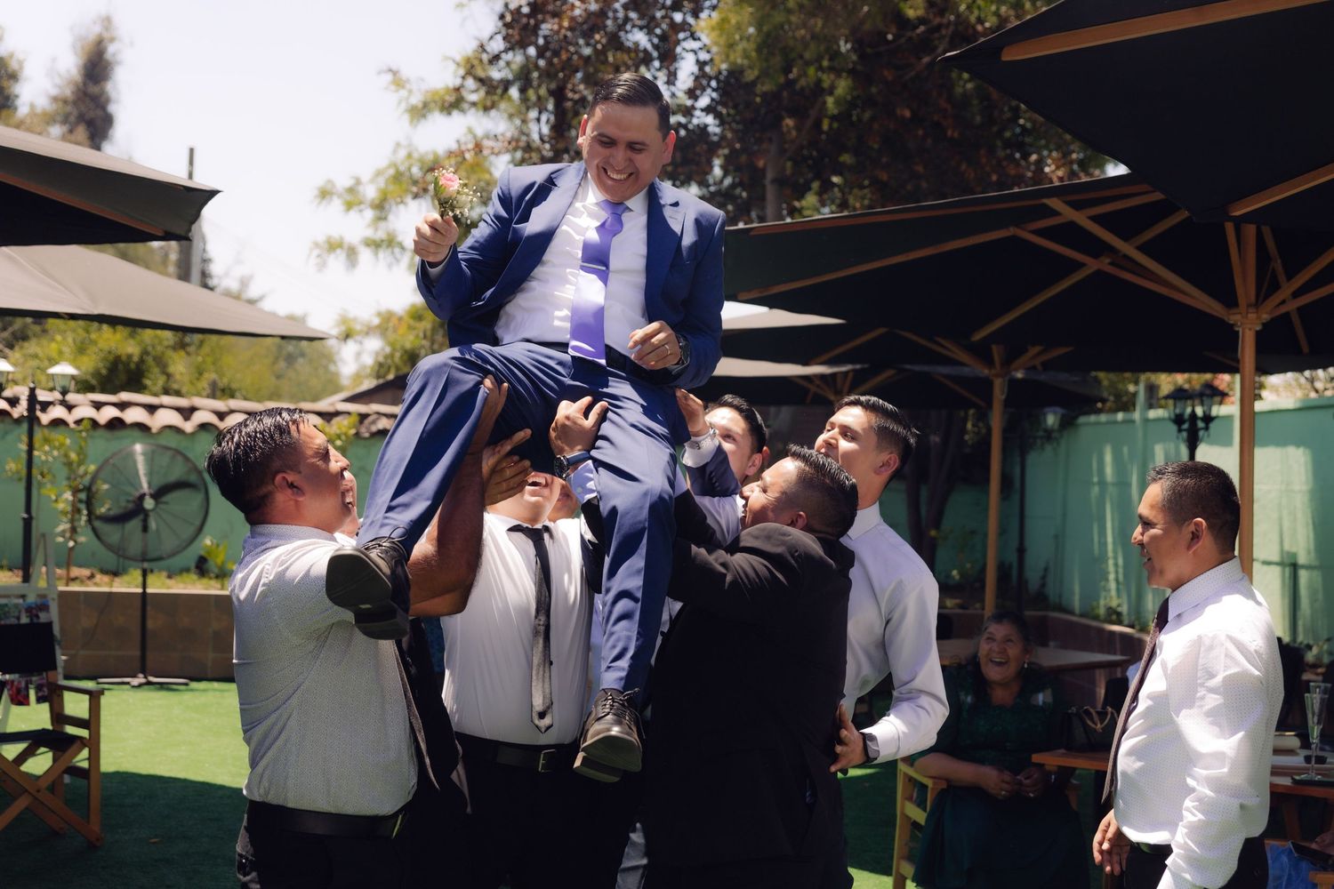 Groomsmen lift groom in blue suit into air during celebratory wedding reception tradition.