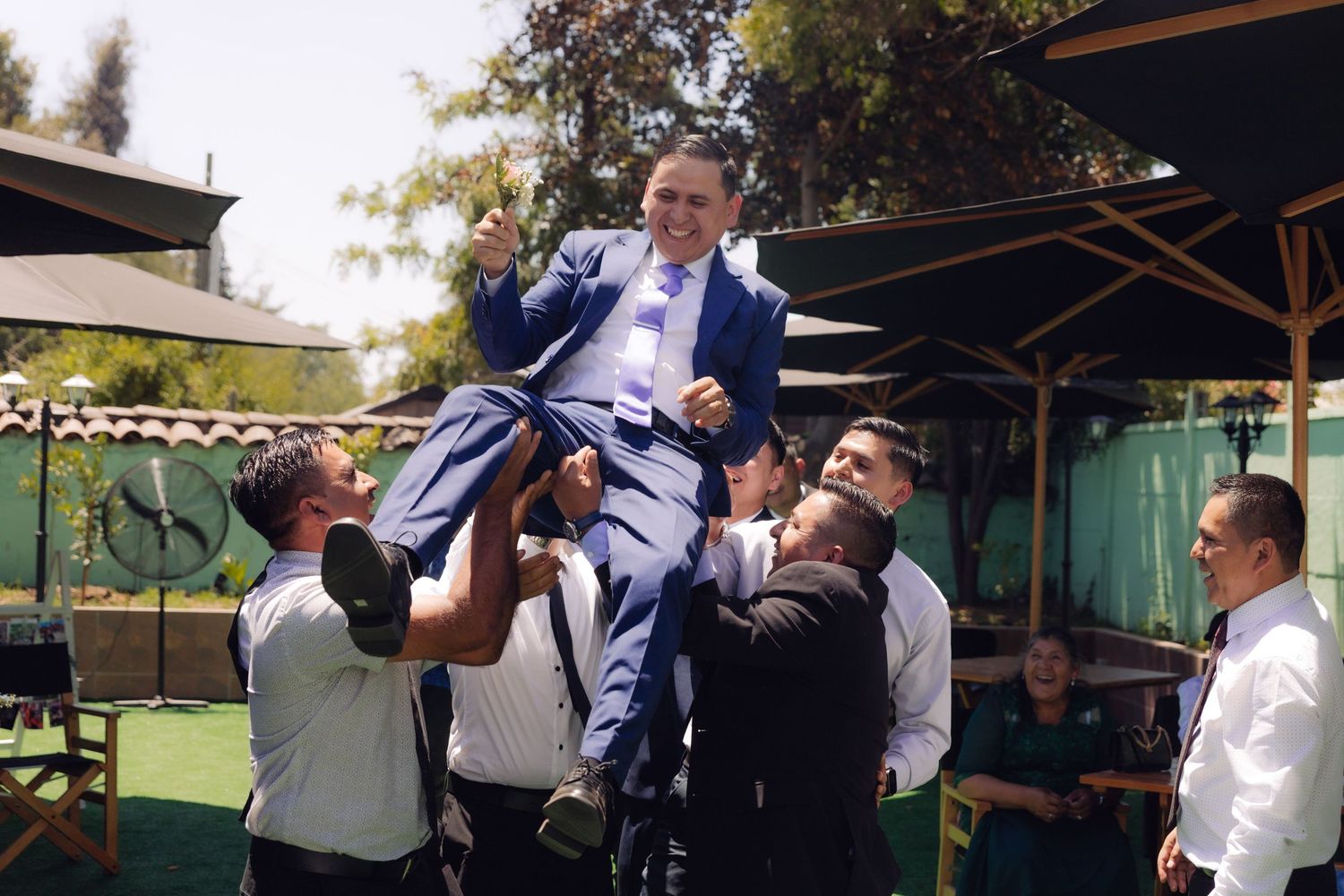 Smiling groom in blue suit lifted by groomsmen during joyful outdoor wedding celebration.