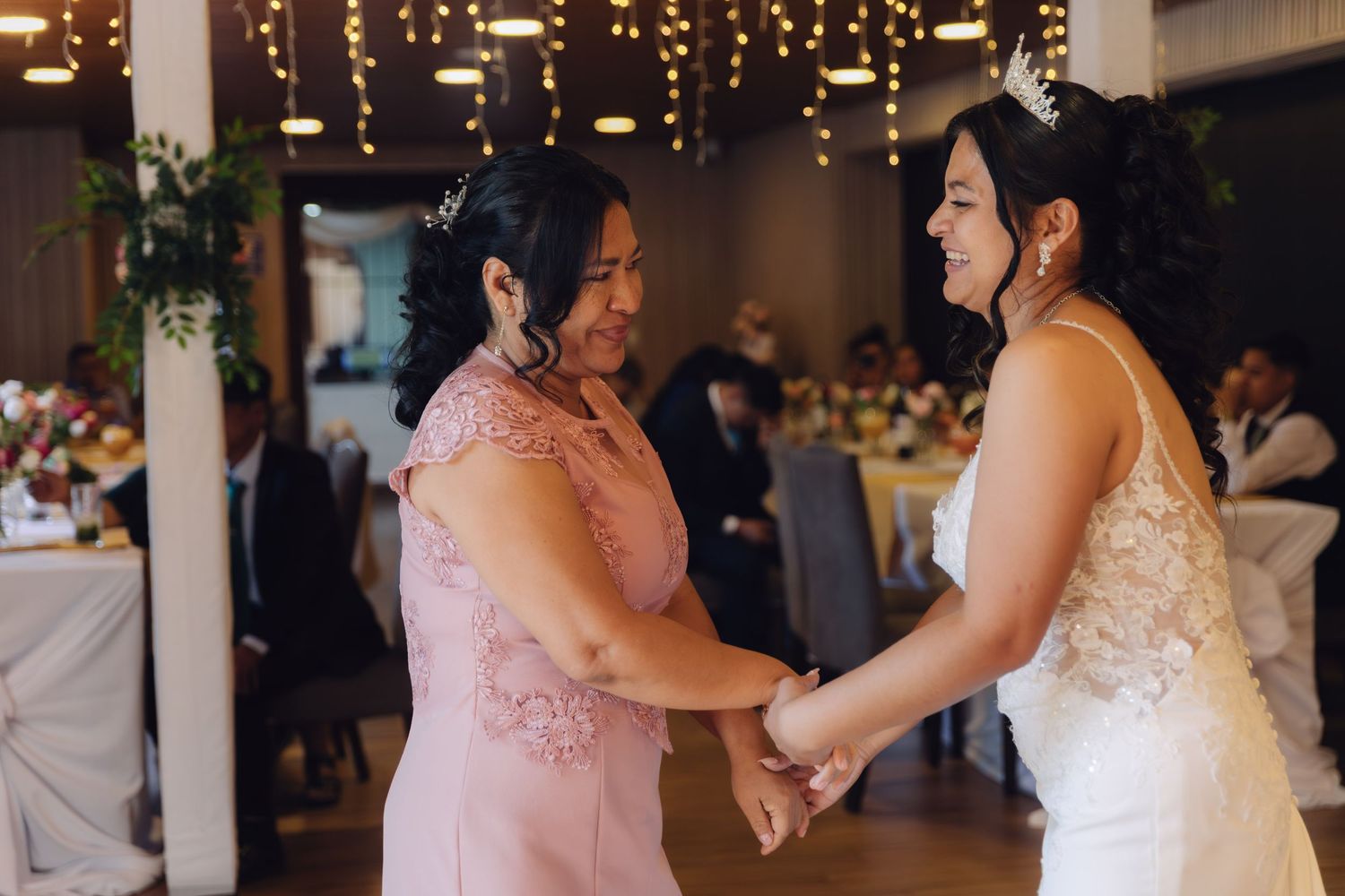 Bride and mother share joyful dance together under string lights at wedding reception.