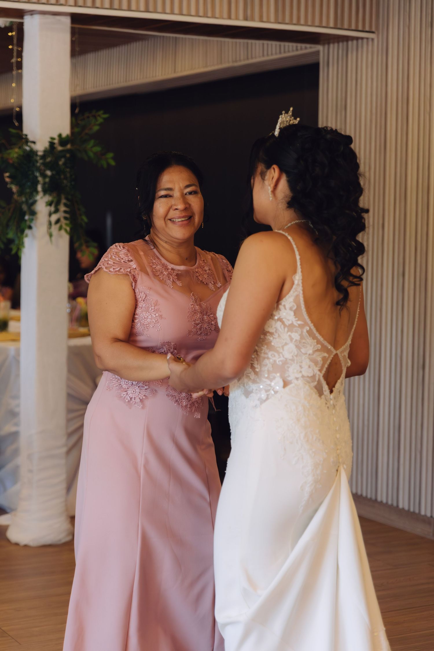 Smiling mother in pink gown holds hands with bride in lace dress at wedding reception.