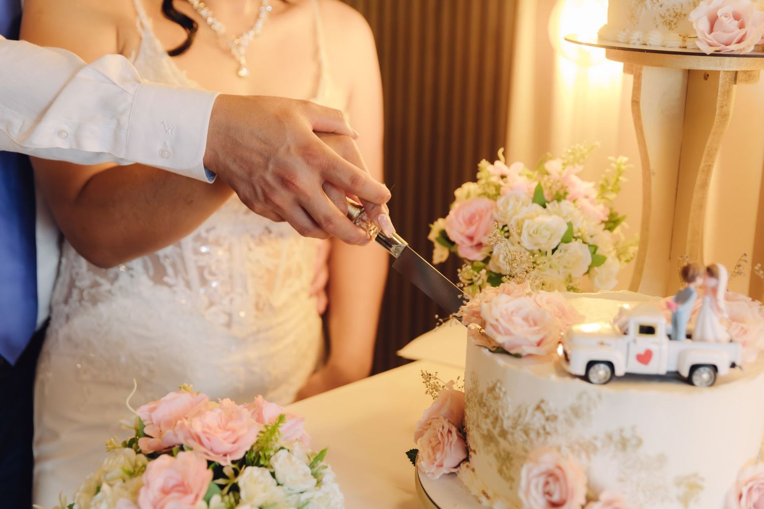 Close-up of bride and groom hands cutting white wedding cake with pink floral decorations.