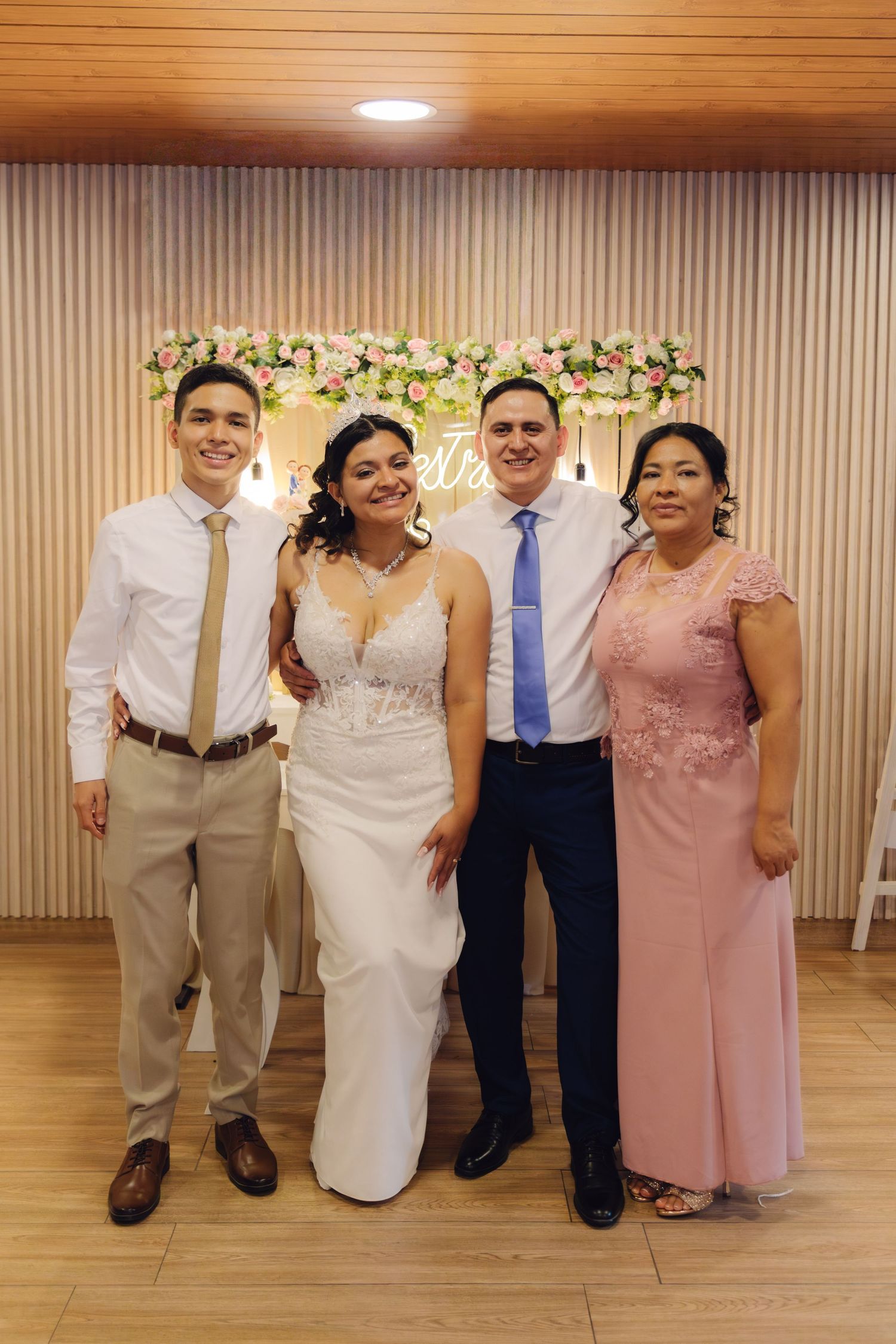 Bride and groom posing with family members in front of floral wedding backdrop display.