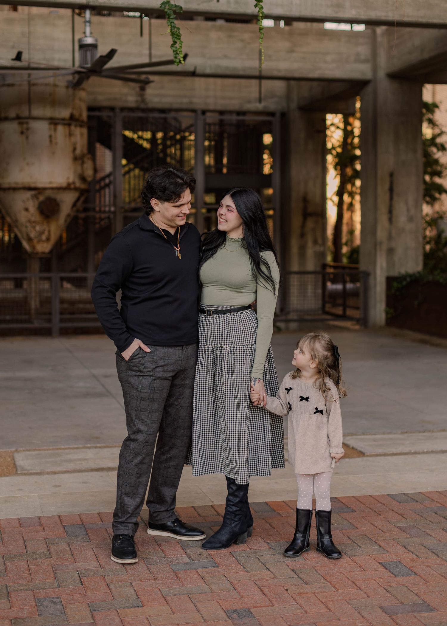 A family of three poses outdoors, parents in dark and green attire with a young girl in a cream dress holding hands.