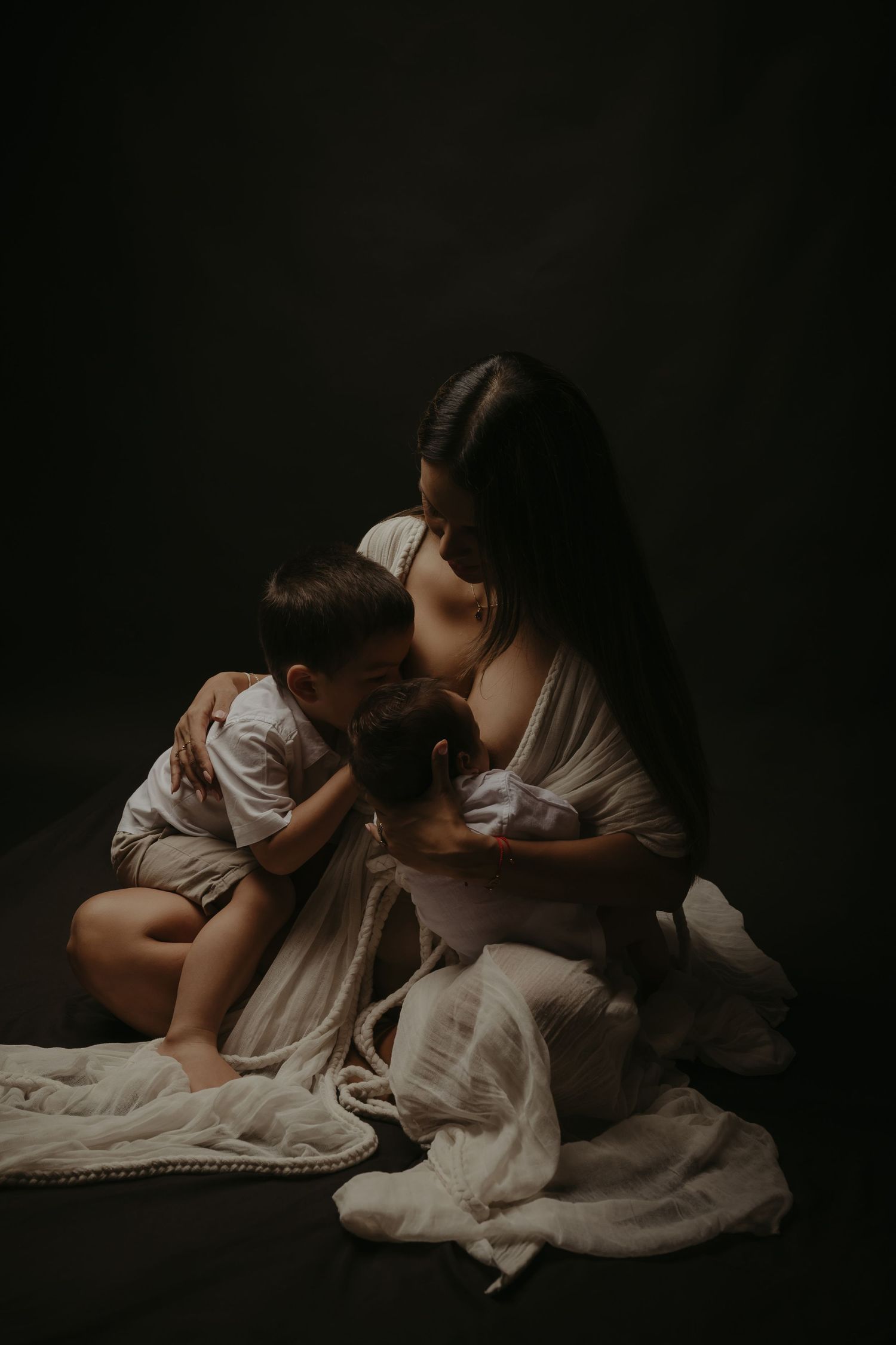 A mother tenderly embraces her two young children while seated on the floor, draped in soft white fabric against a dark background.