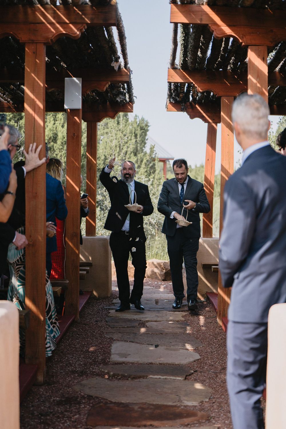 Two groomsmen celebrate walking down the aisle at an outdoor wedding ceremony under a rustic wooden pergola.