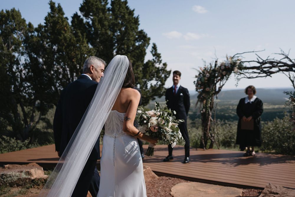 Bride in white gown and veil walks with escort toward groom at rustic outdoor wedding ceremony with tree arch.
