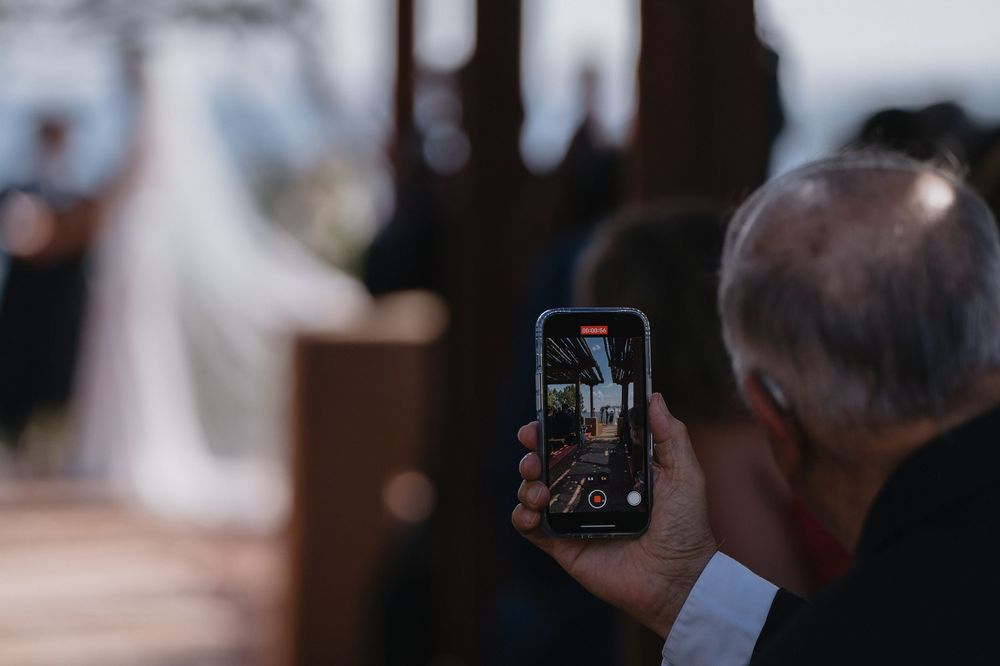A wedding guest captures the ceremony on a smartphone, with the bride visible in the blurred background.