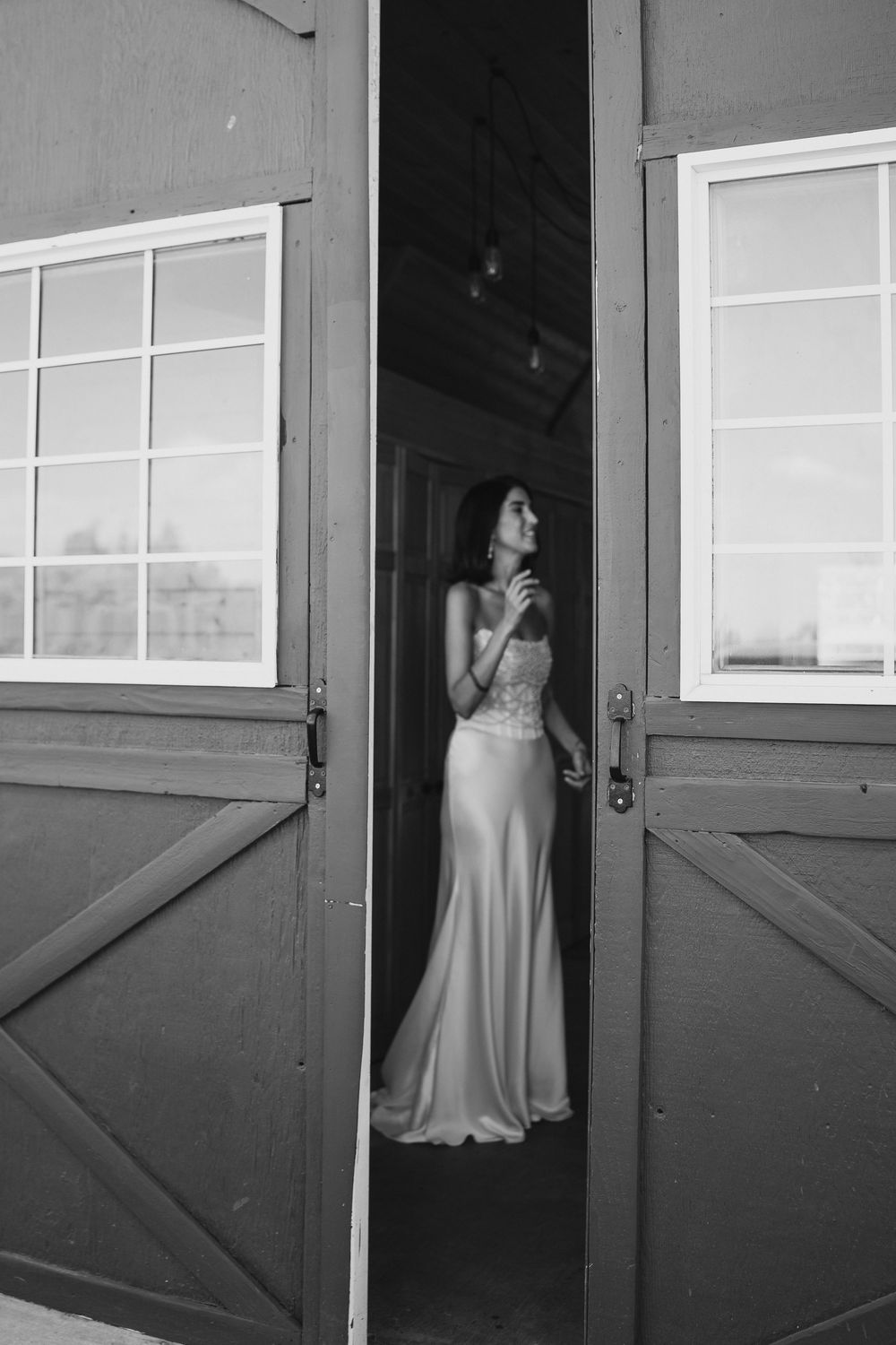 Bride in elegant gown standing in barn doorway, black and white wedding photography.