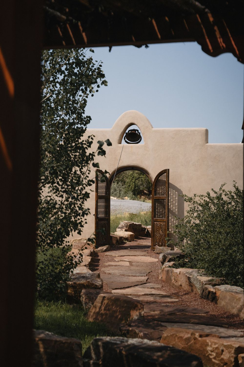 Adobe archway with ornate wooden doors leads to a stone path surrounded by lush trees in a Southwestern landscape.