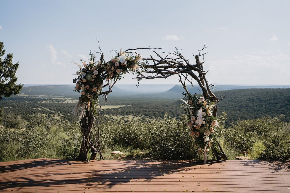 Rustic wedding arch made of twisted branches adorned with flowers on a wooden deck overlooking lush green hills.