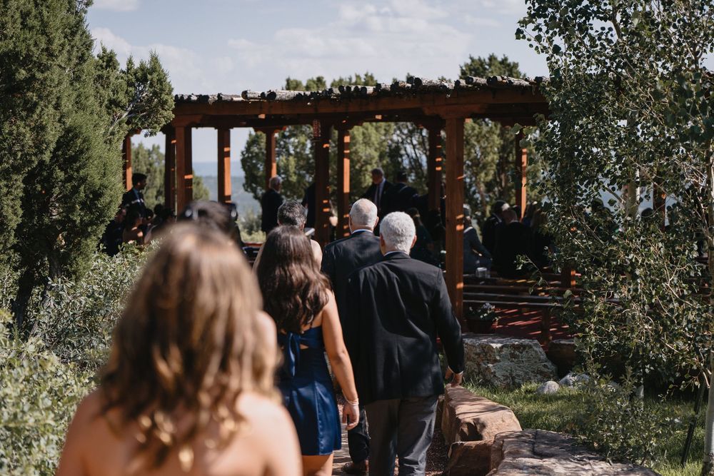 Guests walk along a rustic path toward a wooden pergola ceremony space surrounded by lush greenery outdoors.