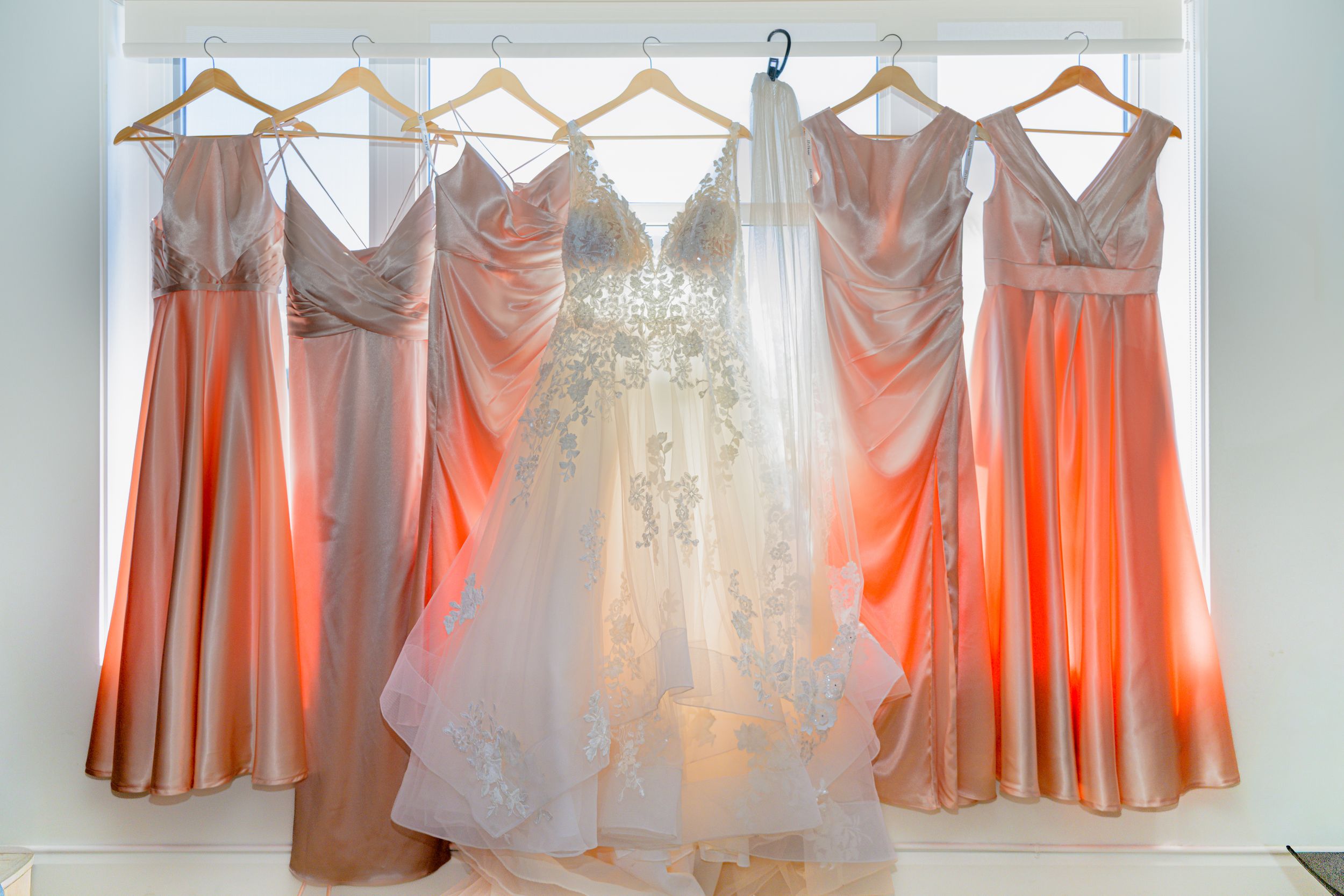 Robe de mariée blanche en dentelle suspendue devant une fenêtre à côté des robes de demoiselles d'honneur roses.