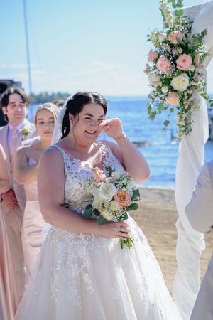 Mariée en robe de dentelle qui rit aux éclats et essuie une larme de joie en tenant son bouquet de fleurs sur la plage de l'Auberge des Îles