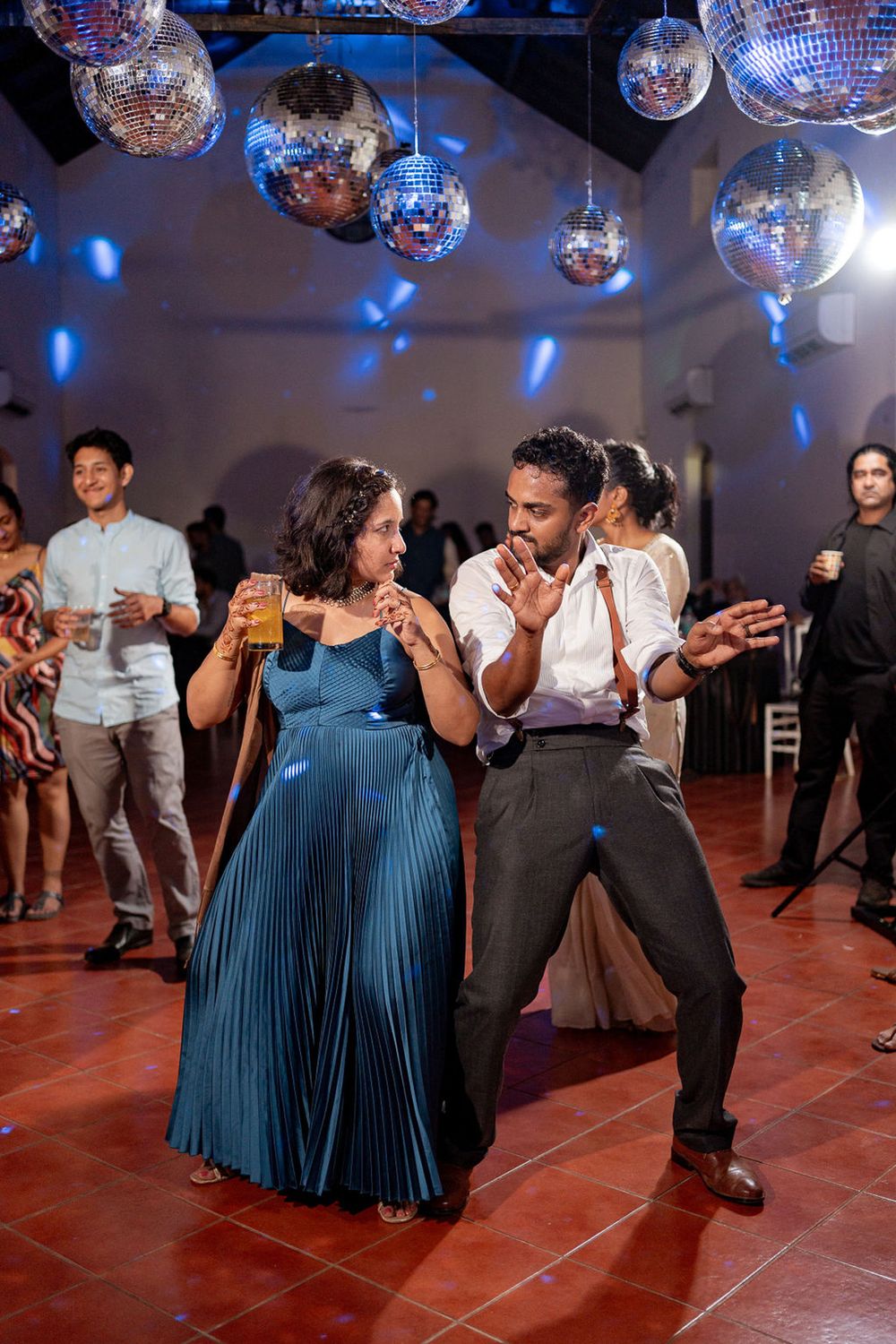Couple dancing joyfully at a party with disco balls overhead, guests watching in a warmly lit venue.