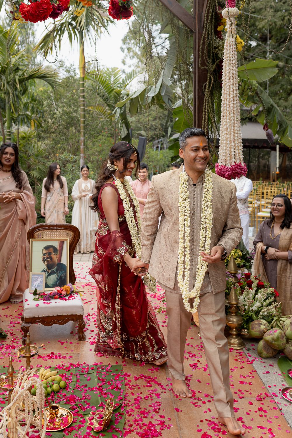 Bride in red lehenga and groom in beige sherwani walk together at an outdoor Indian wedding ceremony with floral decorations.