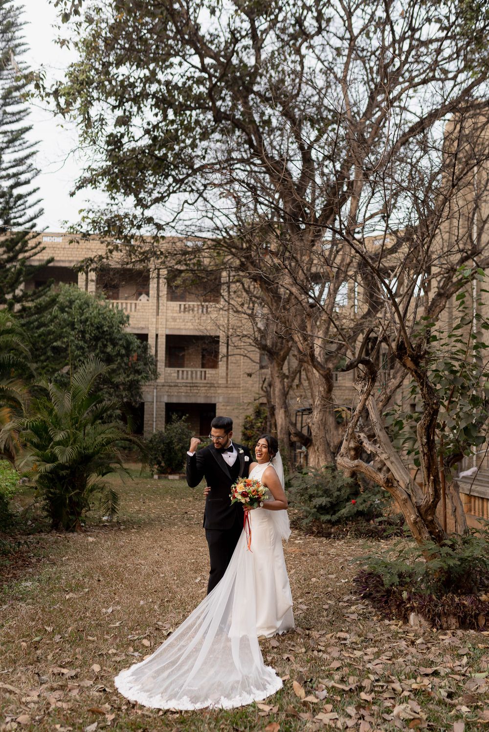 Bride and groom pose in garden of historic stone mansion, bride in elegant white gown with long train holding bouquet.
