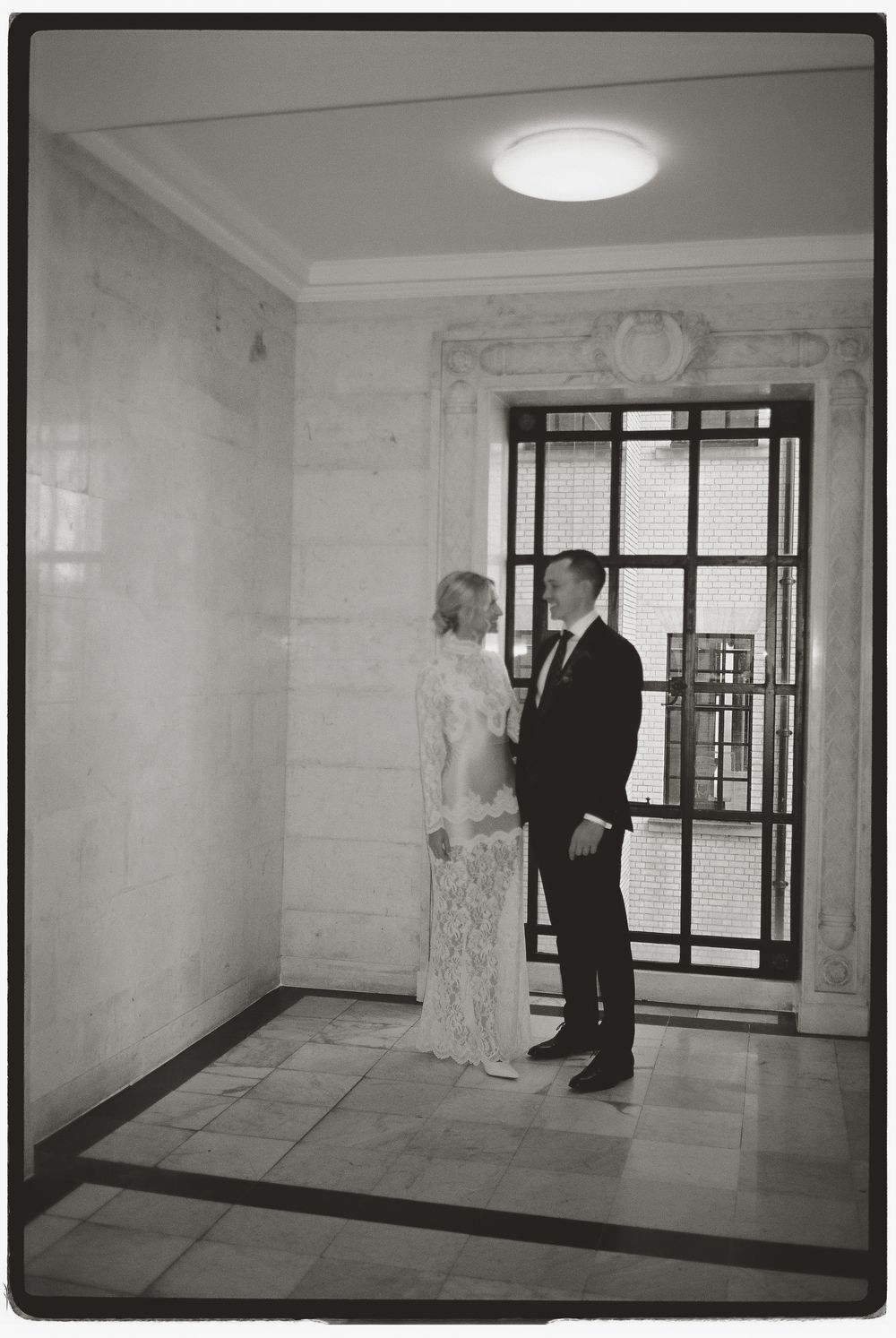 Black and white photo of a bride and groom sharing a romantic moment in an elegant marble hallway.