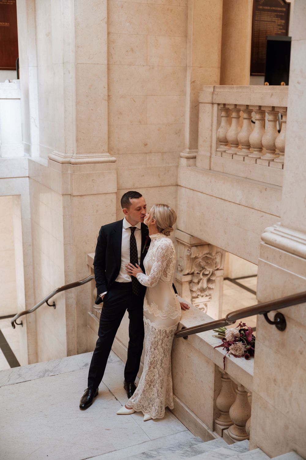 Bride and groom share a romantic moment on marble stairs in an elegant venue, bride wearing lace gown, groom in dark suit.